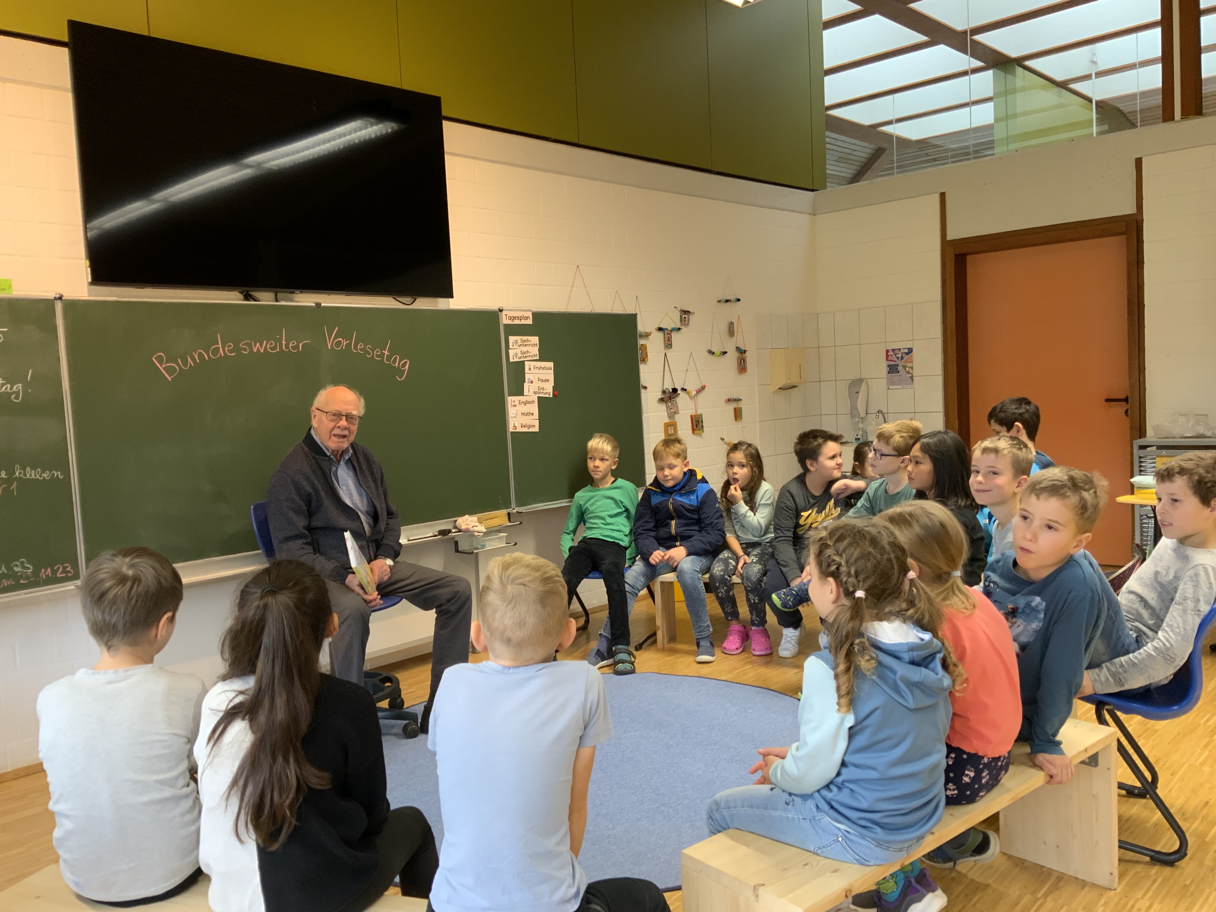 Ein Lehrer liest im Stuhlkreis den Kindern etwas aus einem Buch